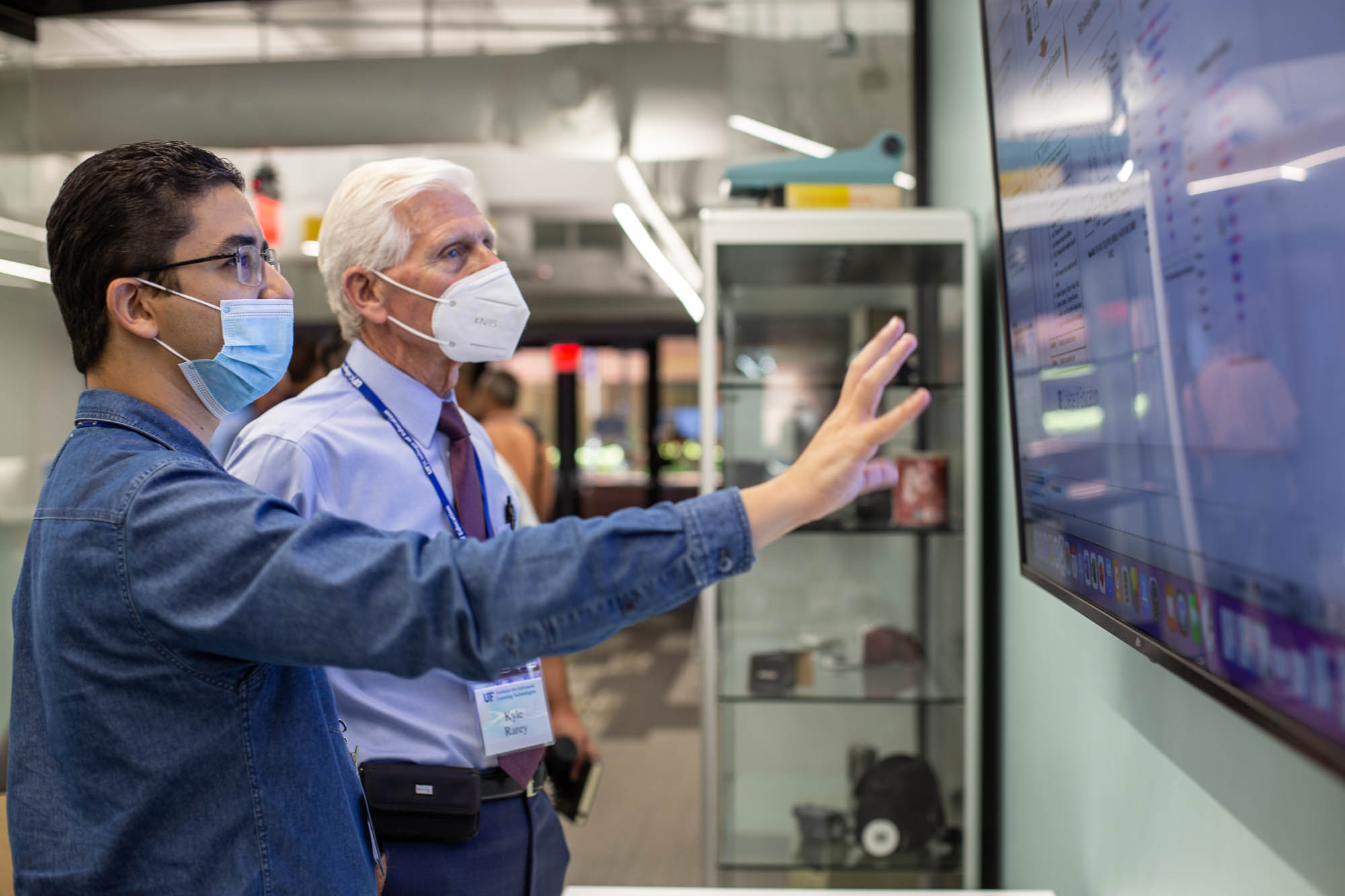 Two people are standing and looking intently at information on a large screen on the wall. The one towards the front has their hand out and explains the information to the other.