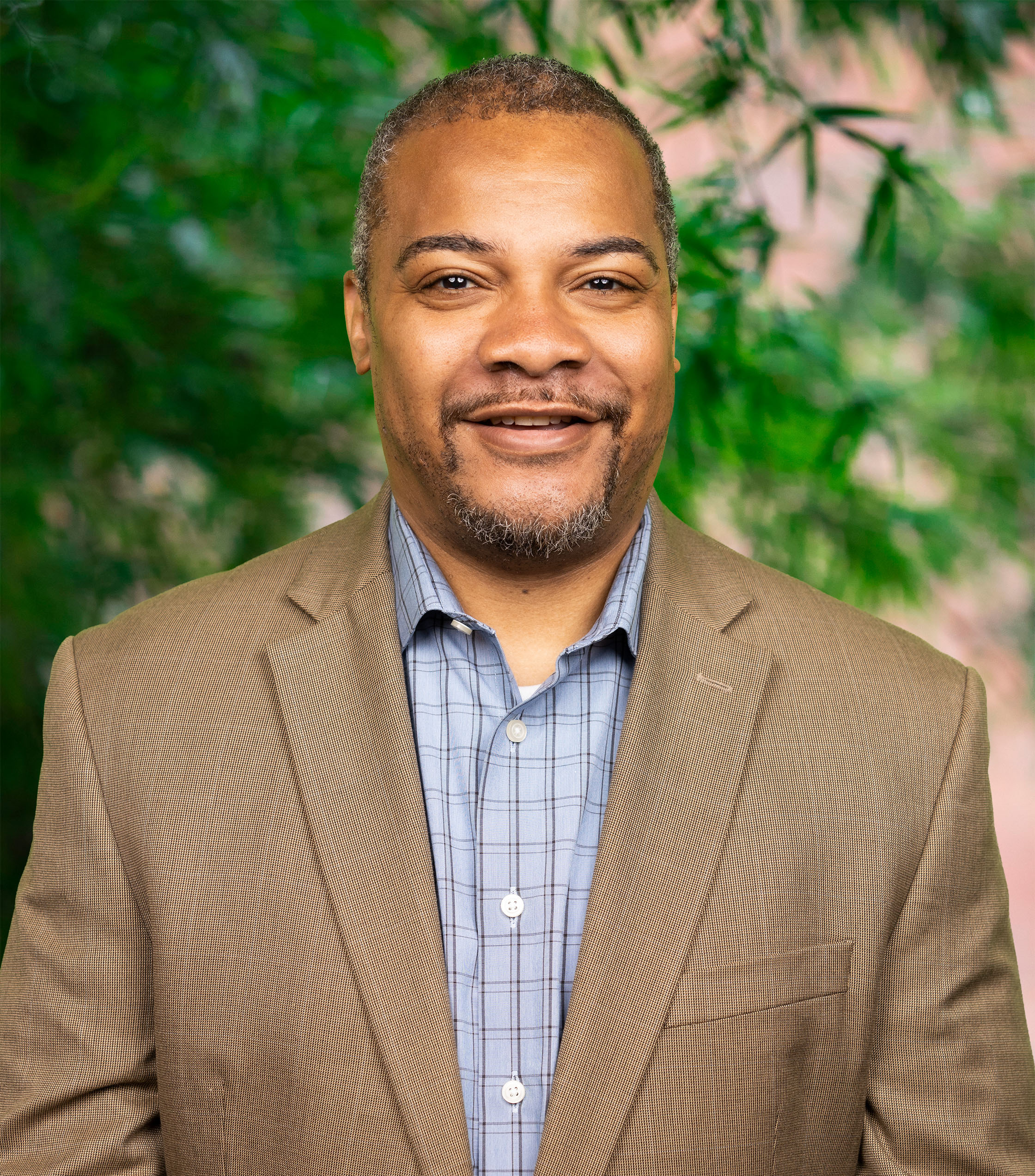 Rob Moore is smiling with trees behind him. He is wearing a brown blazer with a blue button up shirt.